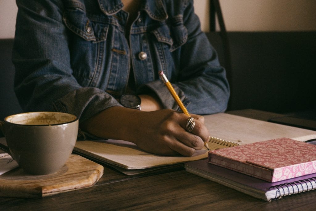 person writing in notepad with coffee and books on table